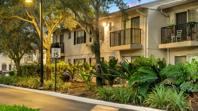 Apartment buildings with balconies and lush landscaping, illuminated by a street light at dusk. A 15 mph speed limit sign.