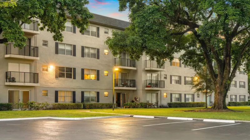 Multistory apartment building with balconies, illuminated at dusk, surrounded by lush trees and a parking lot.