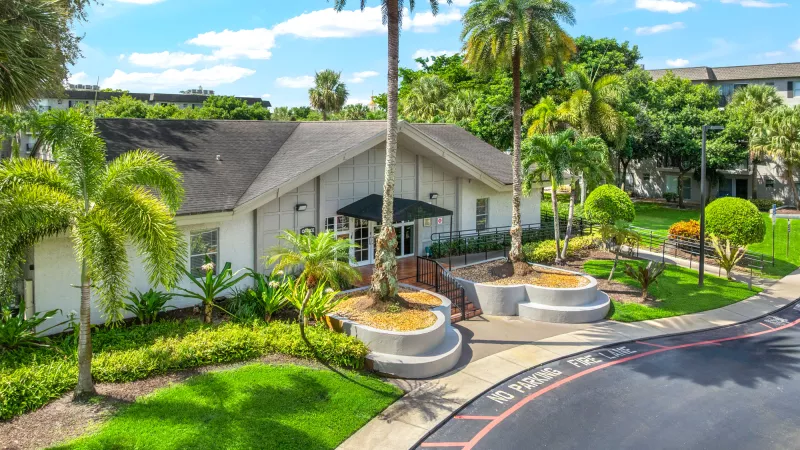 White building with dark roof, surrounded by green grass and tall palm trees, with a paved road curving in front.