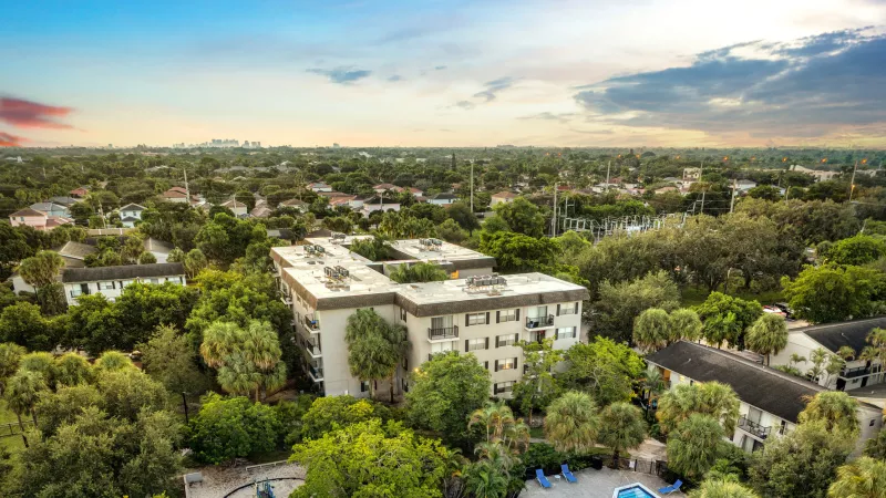 Aerial photograph of residential complex with a large building, blue pool, and lush trees under a colorful sunset sky.