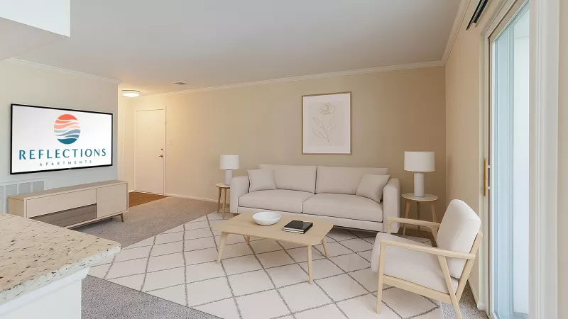 Bright, neutral-toned apartment living room with a beige sofa, matching armchair, and light wood coffee table on a patterned area rug. A TV console with a screen displaying the “Reflections Apartments” logo sits against the left wall, with a framed minimalist floral print above the sofa. Sliding glass doors on the right bring in natural light, and the space opens to a kitchen area in the foreground.