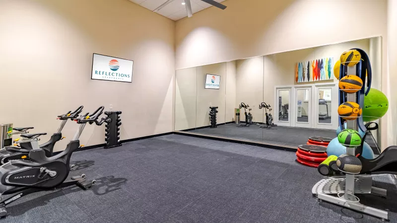 Spacious apartment fitness studio with mirrored walls, stationary bikes, free weights, and colorful medicine balls. A wall-mounted screen displays the Reflections Apartments logo, and the room features high ceilings, a ceiling fan, and dark flooring.