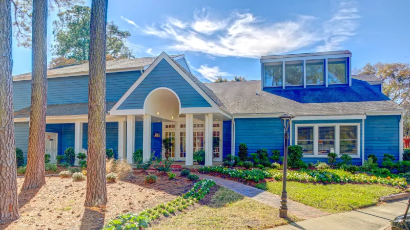 Exterior view of a blue clubhouse building at Reflections Apartments with white columns, landscaped gardens, and a curved walkway leading to the entrance. Tall trees frame the building under a bright blue sky with wispy clouds.