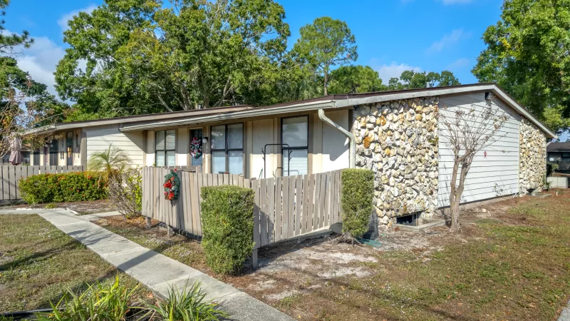 One-story apartment building with stone and siding, a wreath on a wooden fence, sidewalk, and trees.