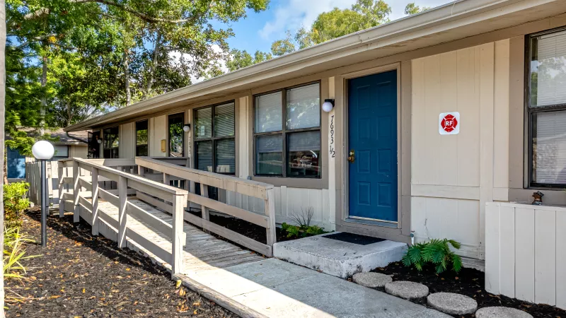 Beige building with a wooden accessibility ramp on the left, multiple windows, and a vibrant blue entrance door on the right.