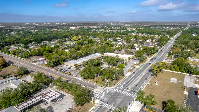 Aerial photograph of a suburban intersection with cars, flanked by numerous green trees and buildings under a blue sky.
