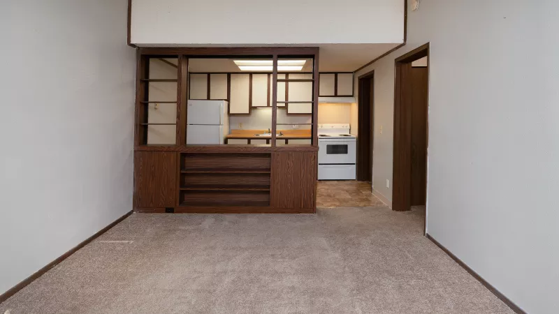 Empty room with beige carpet and white walls, looking into a kitchen with white cabinets through a wooden partition.