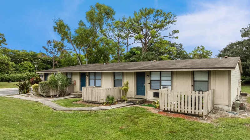 Two beige single-story buildings with blue doors and small fences, a grassy lawn, and trees under a blue sky.