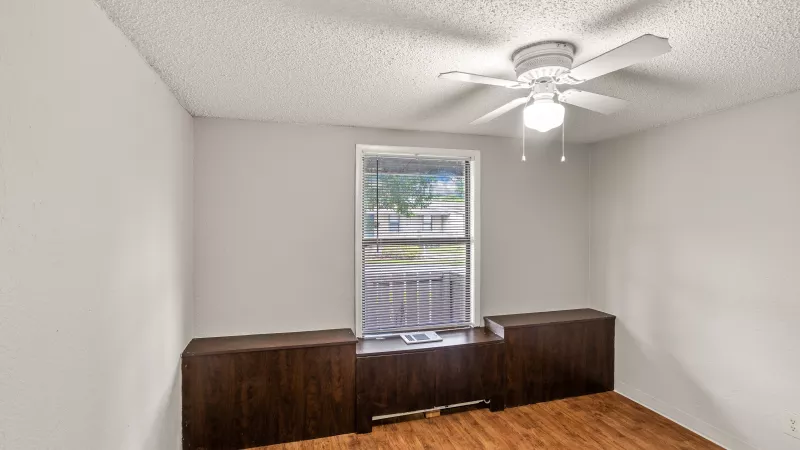 Empty room with white walls, wood floor, window above dark wood built-in storage, and a white ceiling fan.