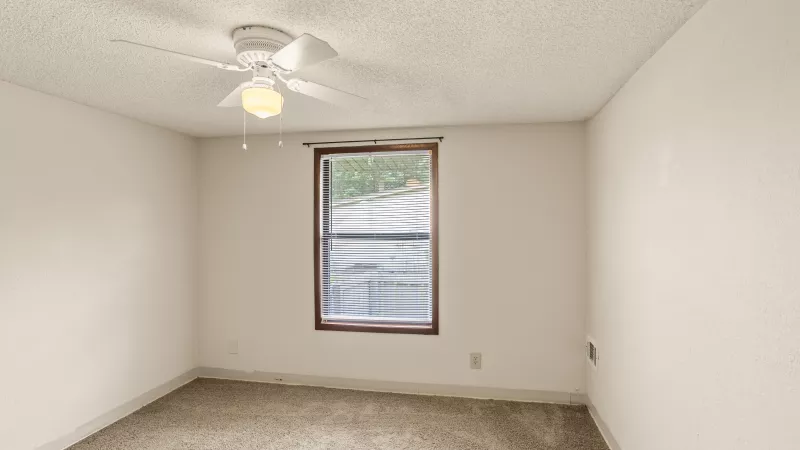 Empty bedroom with white walls, brown carpet, a window with blinds, and a ceiling fan with light.
