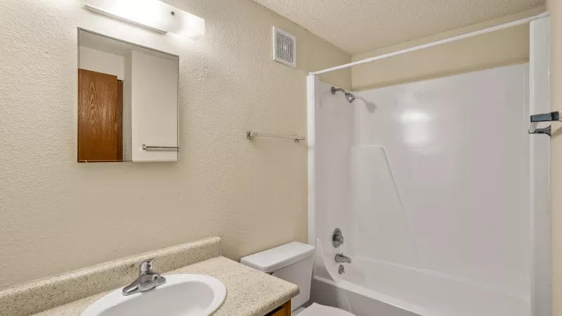 Apartment bathroom with beige walls, a white tub, toilet, and a wood vanity with a white sink and mirror.