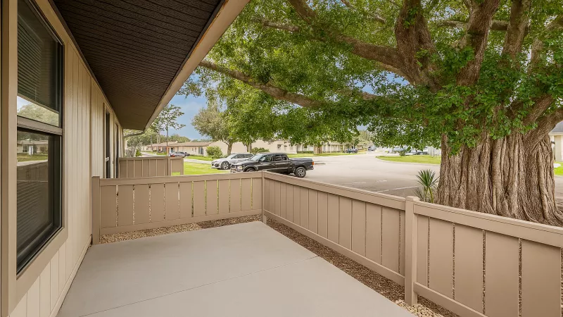 Patio with beige siding, concrete floor, and privacy fence. A large, mature tree stands over a road with parked cars.