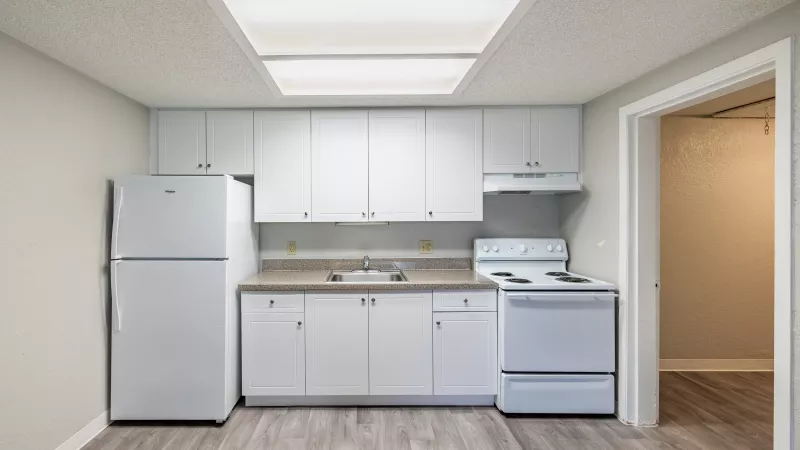 Empty kitchen with white cabinets, light countertop, white refrigerator, electric stove, wood-look floor, and bright ceiling light.