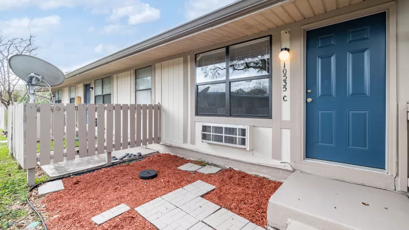 Apartment exterior with a blue front door, beige siding, red mulch patio, and a small AC unit below a window.