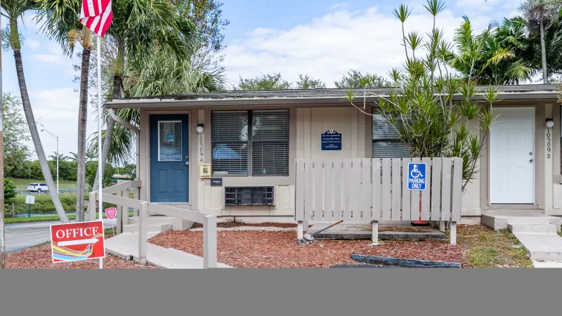 Light brown single-story building with a blue door, an American flag, and a handicap sign, surrounded by mulch and trees.