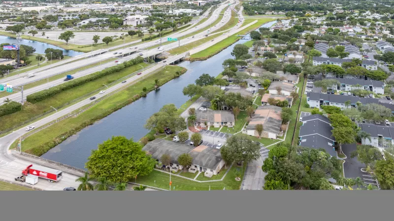 Aerial photograph of a multi-lane highway, a parallel canal, and residential buildings amidst green trees under a clear sky.