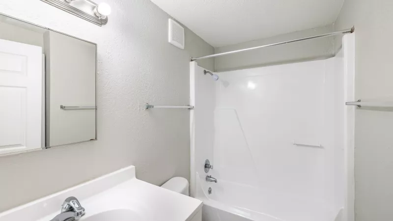 Modern bathroom with white vanity, sink, mirror, toilet, and bathtub/shower unit. Light gray walls.