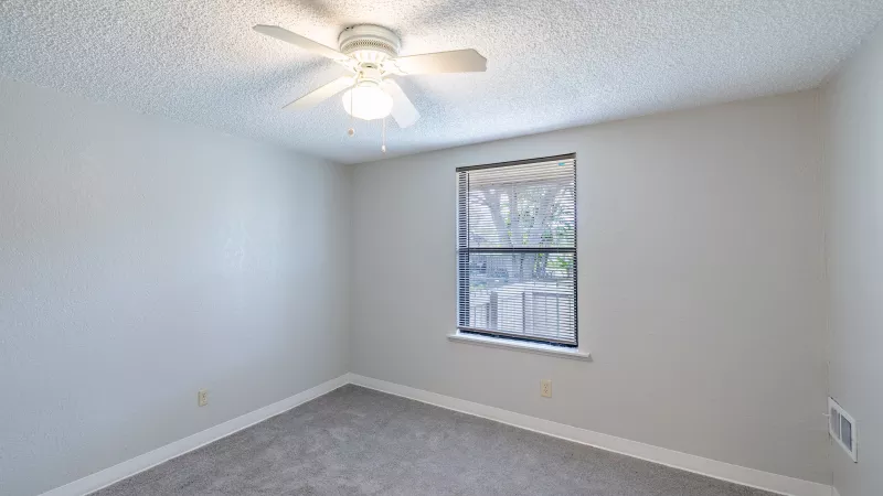 Empty bedroom with light gray walls, gray carpet, a white ceiling fan, and a single window.