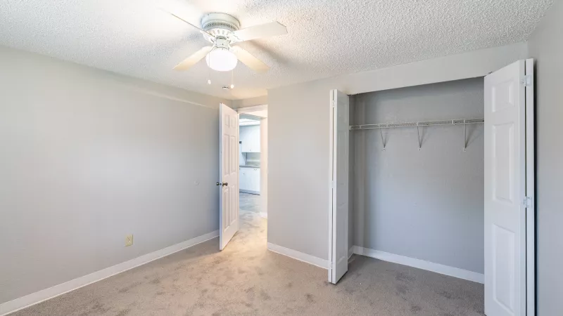 Empty bedroom with light grey walls, carpet, ceiling fan, and open closet with a hanging rod. Doorway to another room.