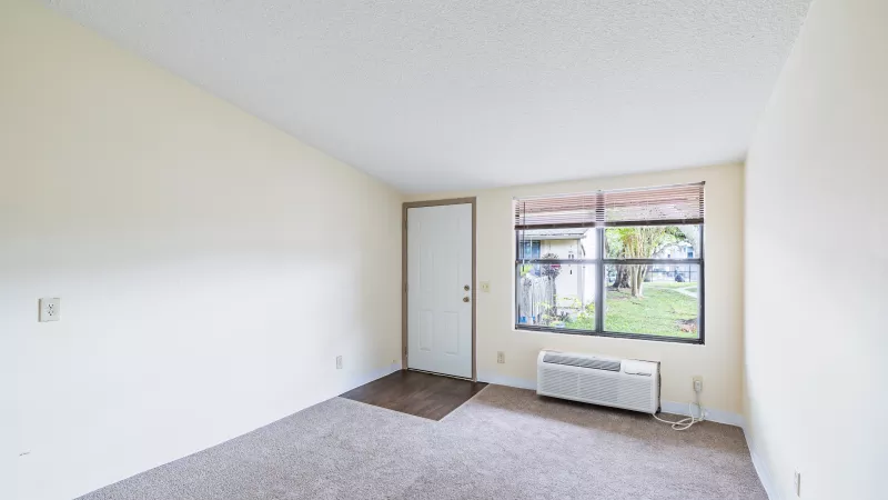 Unfurnished living room with beige walls, brown carpet, a window overlooking trees, and a wall AC unit.
