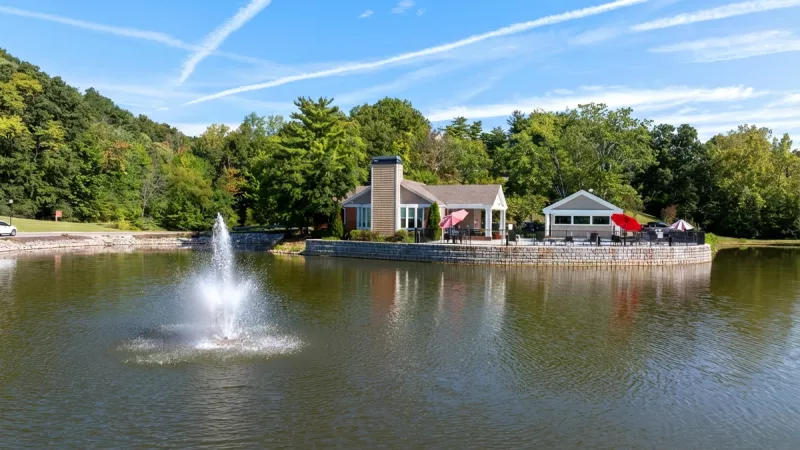 Lakeside clubhouse and patio with red umbrellas, fountain in the water, and green trees under a blue sky.