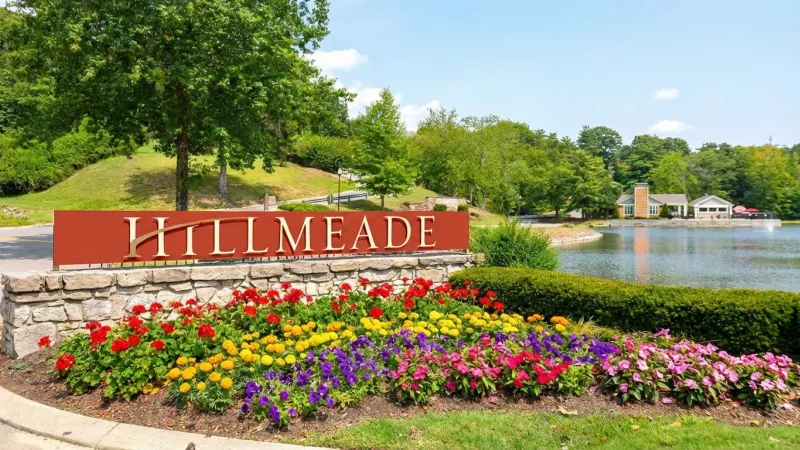"Hillmeade" sign on a stone base, fronted by a vibrant flowerbed. A serene lake, green trees, and houses are in the background.