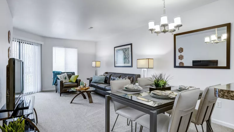 Bright open-plan living room with a dark leather sofa and TV, adjacent to a dining area with a set table and chandelier.