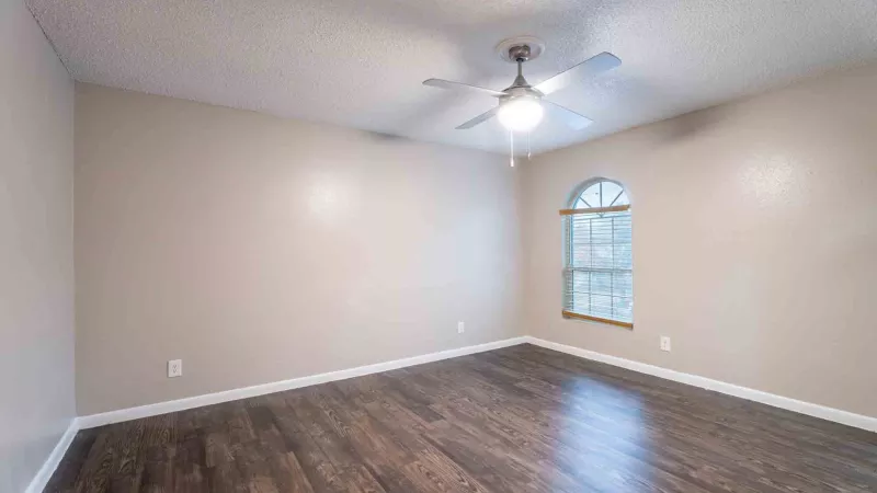 Empty room with light beige walls, dark wood laminate floor, ceiling fan, and an arched window.