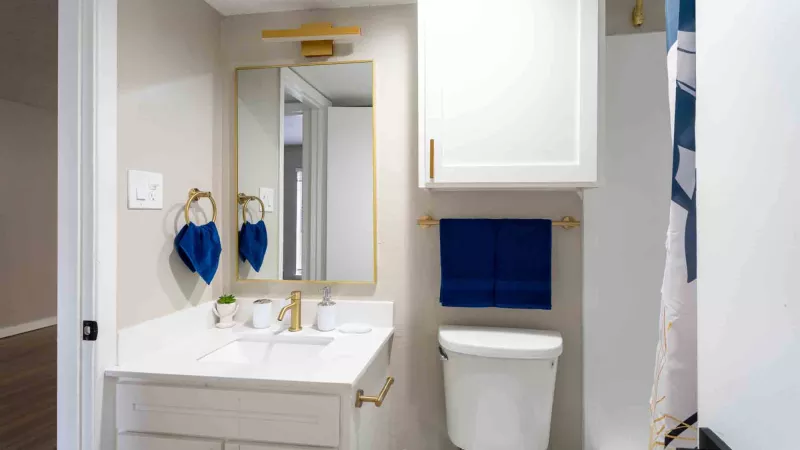 Bathroom with white vanity, gold hardware, blue towels, mirror, toilet, and overhead cabinet.