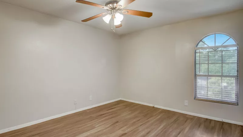 Empty bedroom with light beige walls, wood laminate floor, ceiling fan, and arched window showing trees outside.