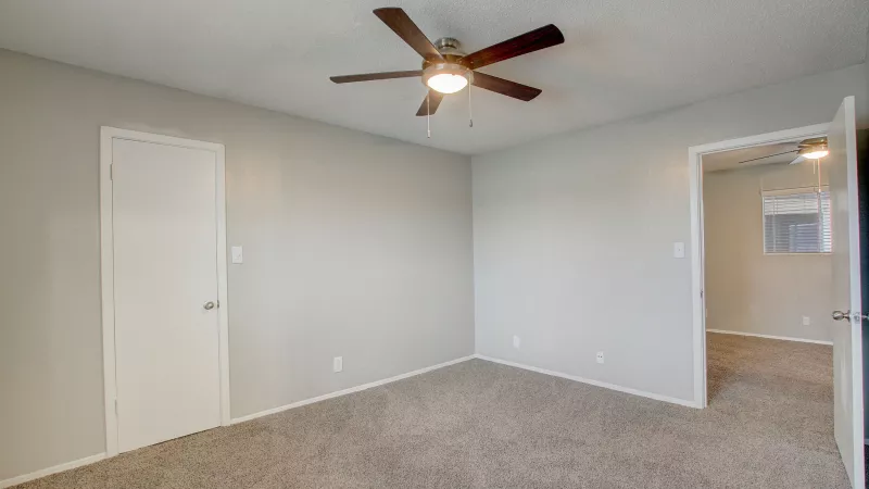 Empty bedroom with light gray walls, brown carpet, ceiling fan, a closed door, and an open doorway to another room.