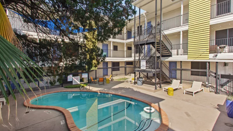 Outdoor swimming pool with lounge chairs in front of a multi-story apartment building, surrounded by green trees.