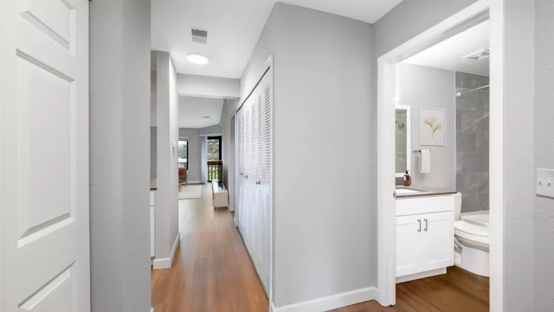 Long hallway with light gray walls, wood floor, and open doorway to a bathroom on the right. White doors line the left and right.
