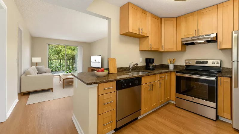 Open-concept kitchen with light wood cabinets, dark counters, stainless appliances, and a living room in background.