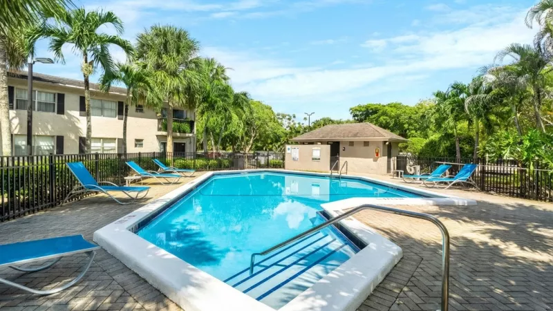 Sunny swimming pool with blue lounge chairs, surrounded by palm trees and apartment buildings.