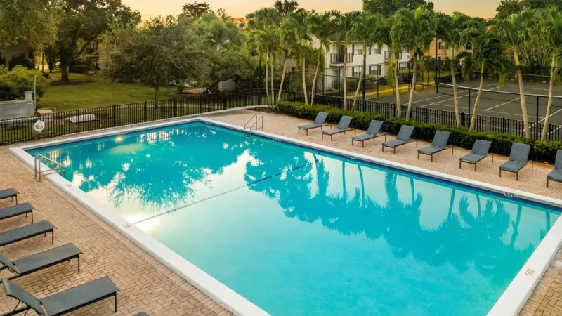 Large rectangular swimming pool with many lounge chairs, surrounded by palm trees and apartment buildings at sunset.
