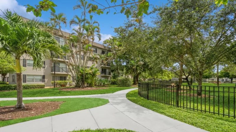 Winding path through a green lawn with palm trees leading to an apartment building under a blue sky.