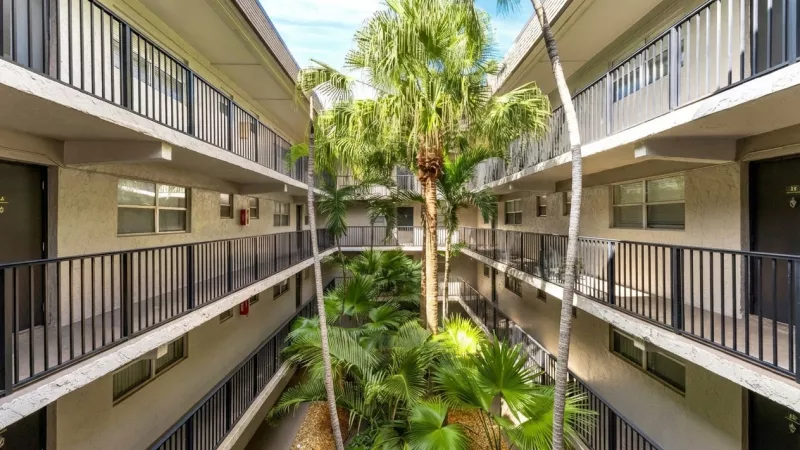 Apartment building courtyard with multiple floors of balconies, black railings, and a lush central garden with palm trees.