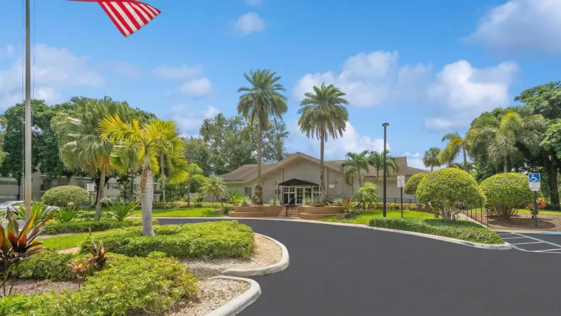 Building entrance with an American flag, palm trees, and manicured landscaping under a blue sky.