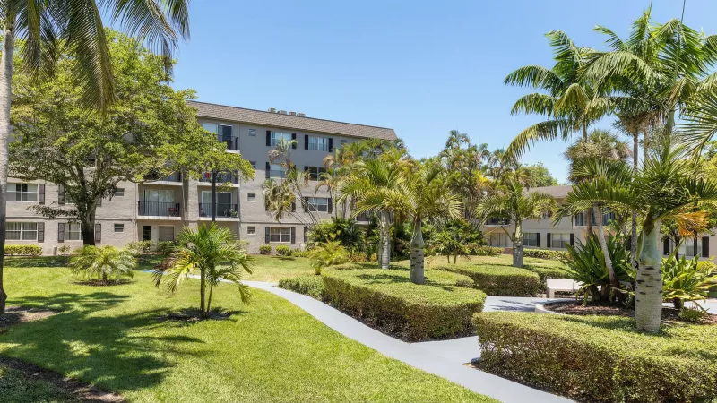 Tropical apartment complex with green lawns, numerous palm trees, and a paved walkway on a sunny day.