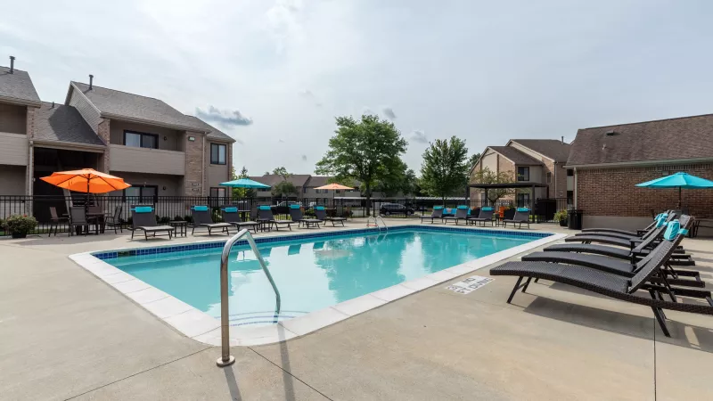 Outdoor swimming pool with blue water, lounge chairs, and colorful umbrellas surrounded by apartment buildings.