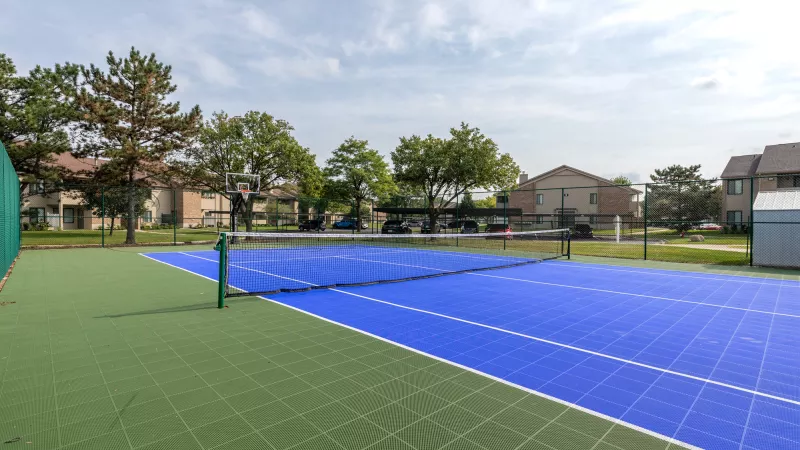 Outdoor tennis court with green and blue surfaces, net, trees, and residential buildings under a cloudy sky.