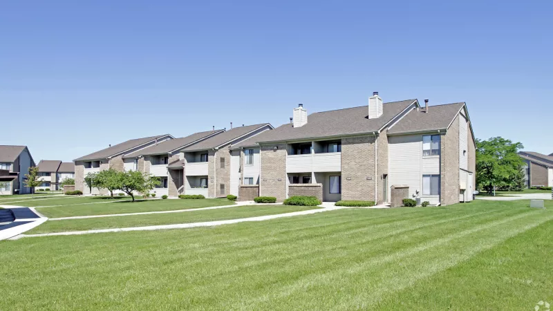 Row of apartment buildings with light siding and brick accents, fronted by a large green lawn under a clear blue sky.