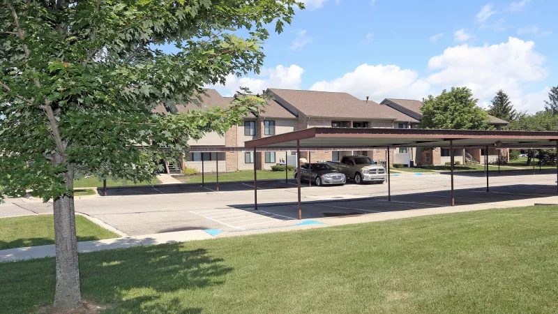 Sunny view of apartment buildings, a parking lot with brown carports over cars, green grass, and trees.