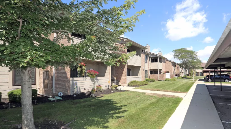 Suburban apartment buildings with brick and siding, green grass, trees, and covered parking under a blue sky.