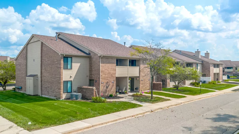 Multiple two-story brick residential buildings with beige siding, green lawns, and a partly cloudy blue sky.