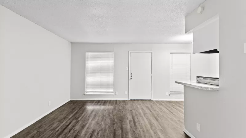 Empty living room with dark wood floors, light walls, two windows with blinds, a white door, and a kitchen counter.