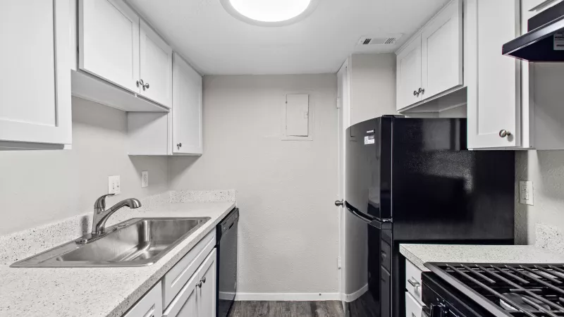 Clean kitchen with white cabinets, light speckled countertops, stainless steel sink, black refrigerator, and dark wood floor.