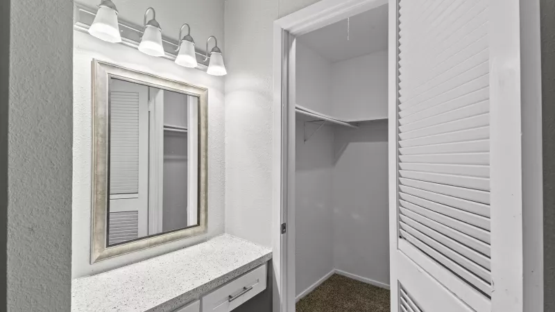 Modern white vanity with mirror, overhead lights, and drawers, beside an open closet with louvered bifold doors and brown carpet.