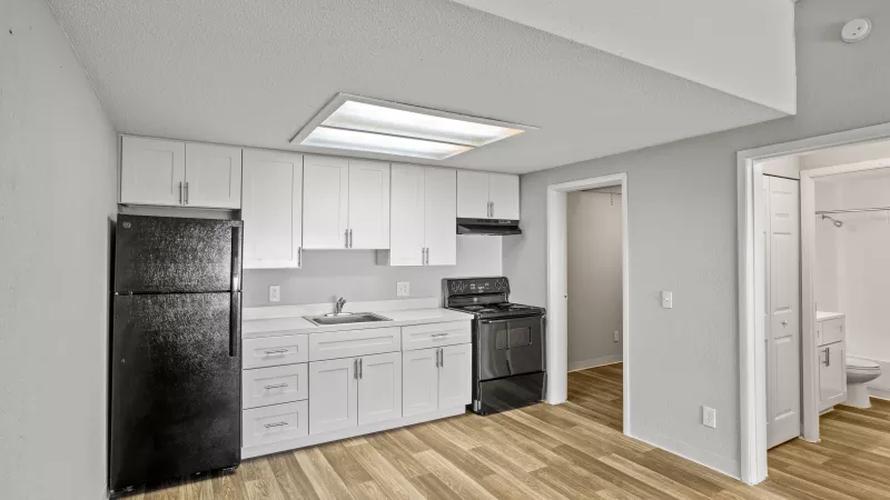 Compact kitchen with white cabinets, black appliances, and wood floors. Bathroom visible through doorway on right.